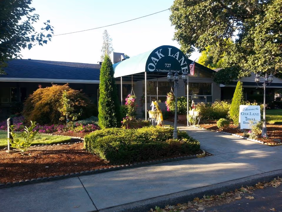 Front exterior view of Oak Lane Assisted Living & Memory Care building with a green awning displaying the name and address. The entrance is surrounded by well-maintained landscaping including bushes, flowers, and trees. A sidewalk leads to the entrance, and there is a sign on the right side welcoming visitors.