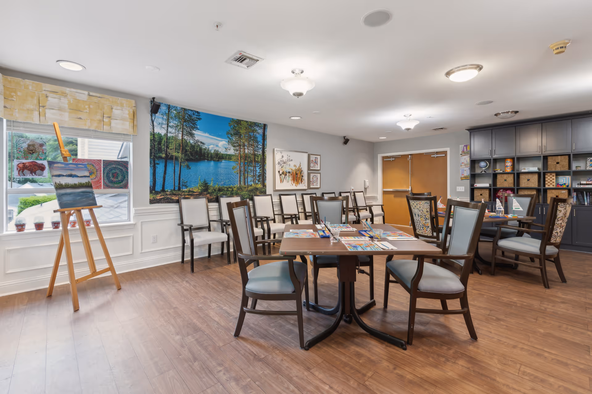 A well-lit activity room in a senior living facility with several tables and chairs arranged for group activities. The tables have coloring supplies and papers on them. There is an easel with paintings near a window, and the walls are decorated with artwork, including a large scenic photograph of a lake and trees. Shelving units with baskets and books are visible in the background.