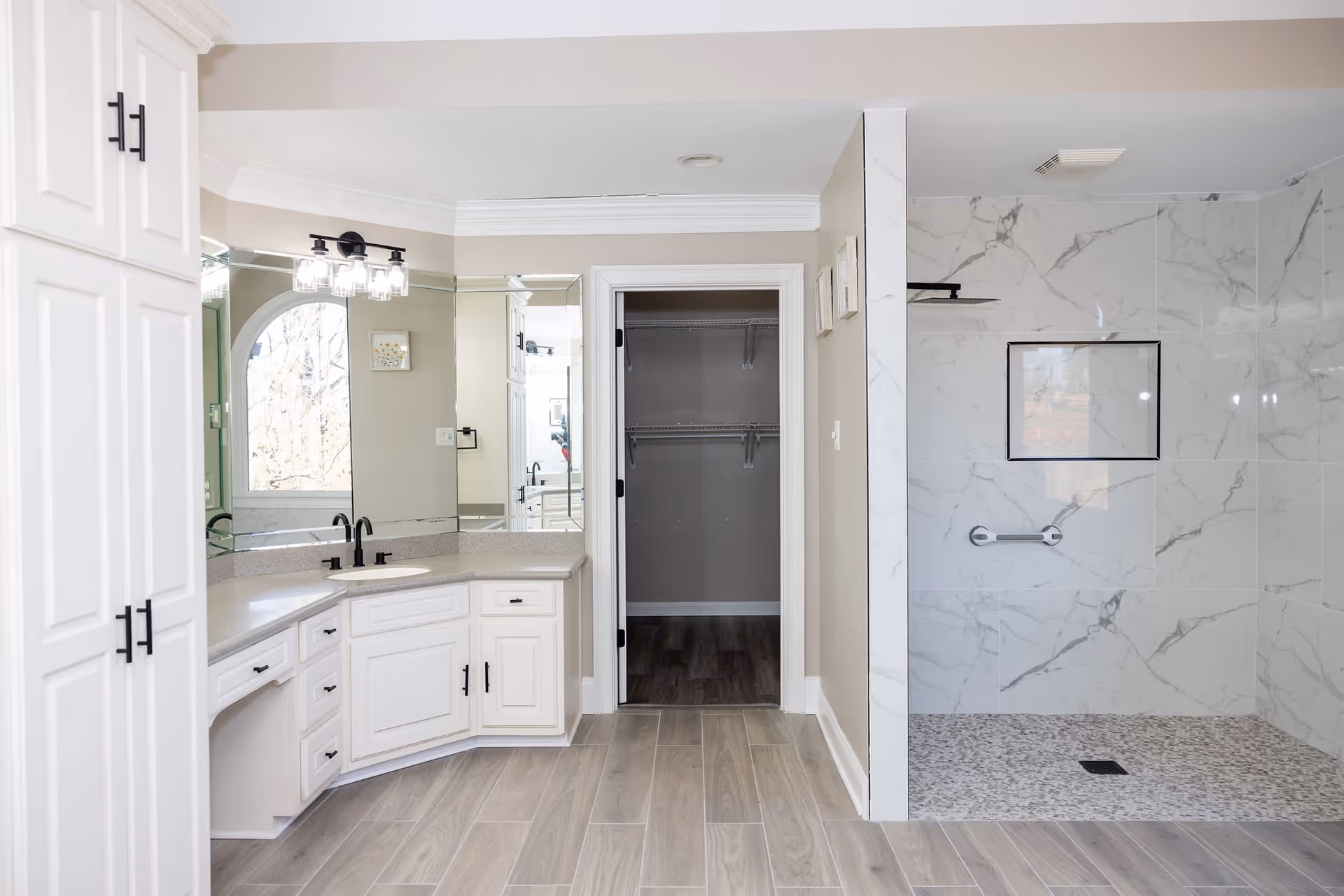 Bright bathroom with white cabinetry, a large mirror, a countertop with a sink and black faucet, a walk-in shower with marble-patterned tiles and a built-in shelf, and an open doorway leading to a closet with wire shelving.
