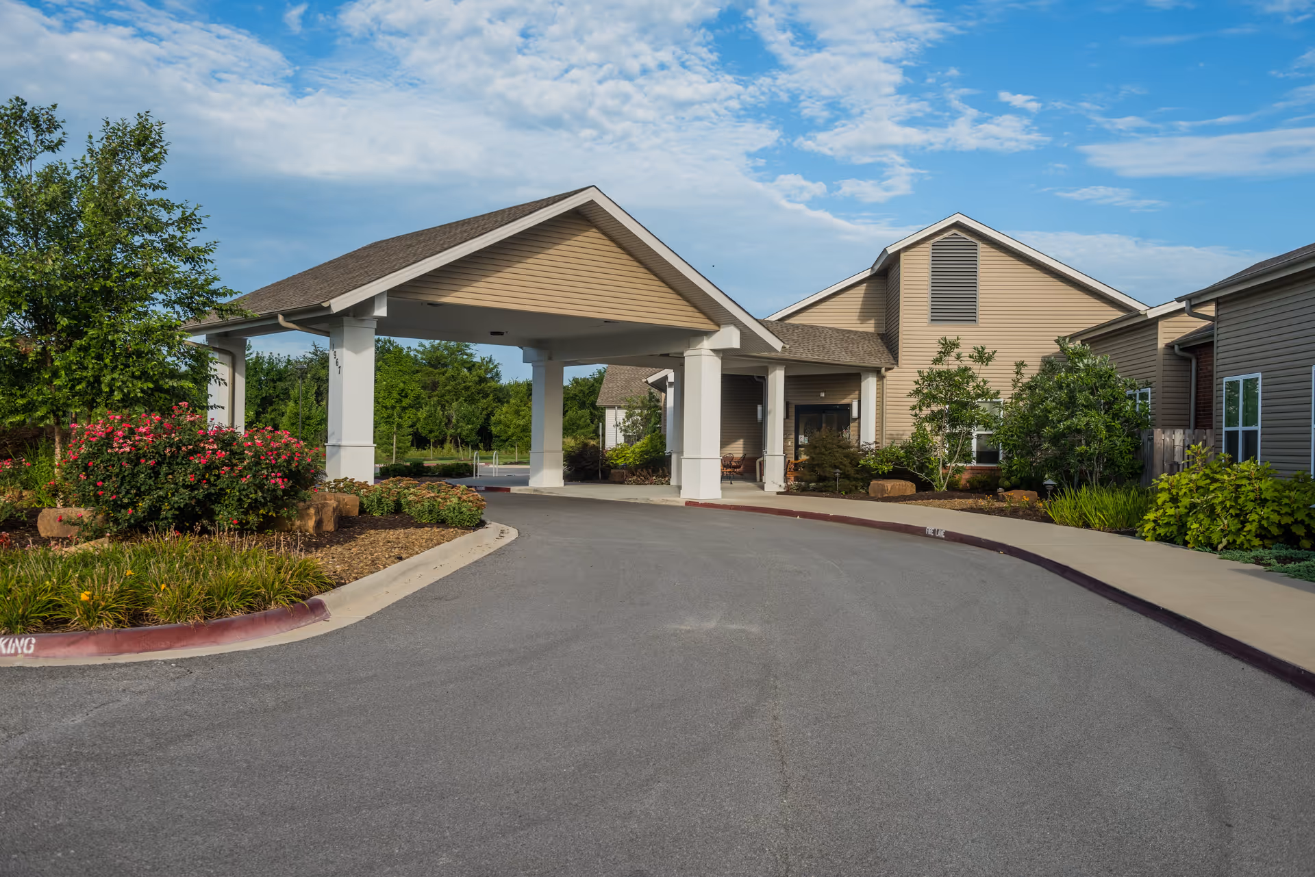 Entrance of Clear Creek Memory Care facility showing a covered drop-off area with beige siding buildings, landscaped bushes, trees, and a clear blue sky with some clouds.