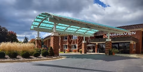 Exterior view of Fairhaven Nursing Home showing the main entrance with a covered drop-off area supported by brick pillars. The building is made of red brick with multiple windows, and there are landscaped bushes and plants around the entrance. The sky is partly cloudy.