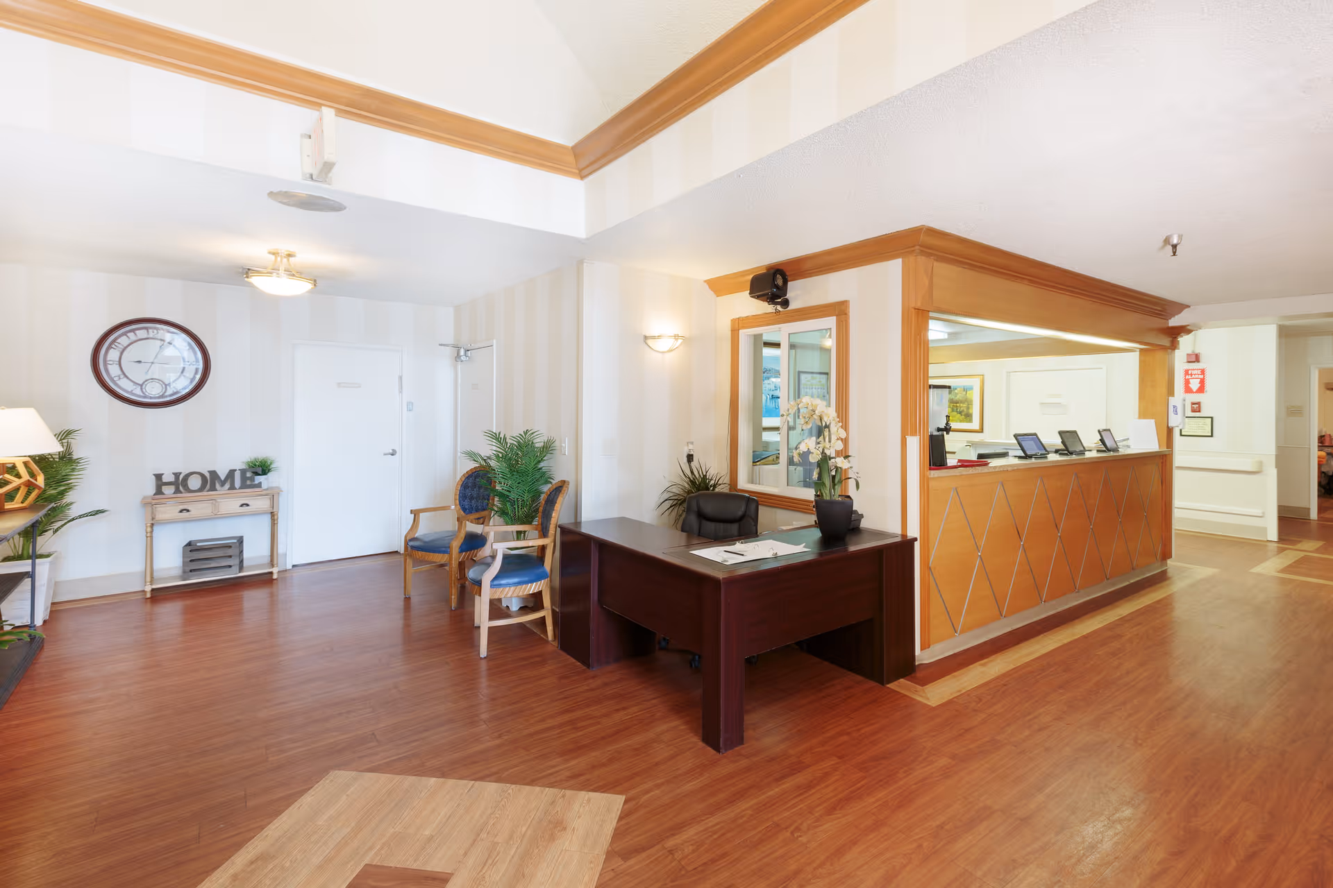 Reception area of a senior living facility with a wooden desk, chairs, potted plants, a wall clock, and a small table with decorative items including a sign that says 'HOME'. The space has wood flooring and light-colored walls with crown molding.