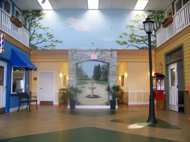 Interior view of a senior living facility hallway designed to resemble an outdoor street scene, featuring a mural of a garden path with a fountain, potted plants, a street lamp, a popcorn machine, and storefront-style doors and windows with awnings.