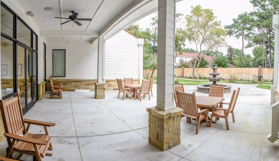 Covered outdoor patio area with wooden tables and chairs, ceiling fans, and a view of a fountain and trees in the background.