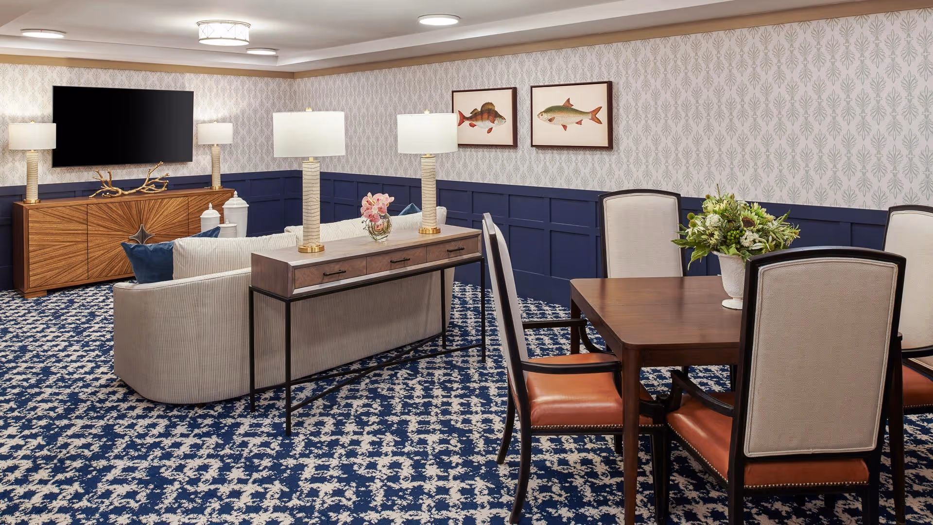A cozy living room and dining area in a senior living facility with patterned blue and white carpet, a curved beige sofa, a wooden console table with two lamps and a flower vase, a wooden dining table with four upholstered chairs, a wooden sideboard with two lamps and decorative items, a wall-mounted flat-screen TV, and two framed fish illustrations on the wall.