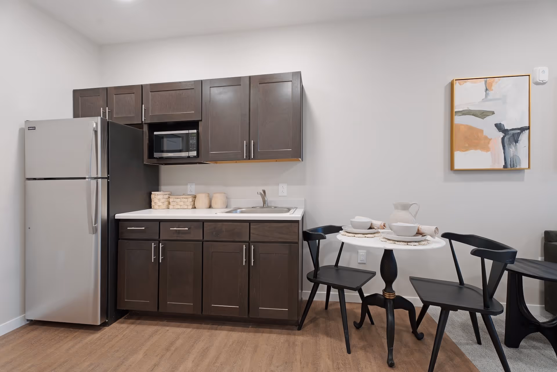 A small kitchen area with dark wood cabinets, a stainless steel refrigerator, a microwave, and a sink. Next to the kitchen is a small round white table set for two with black chairs. A modern abstract painting hangs on the wall above the table.