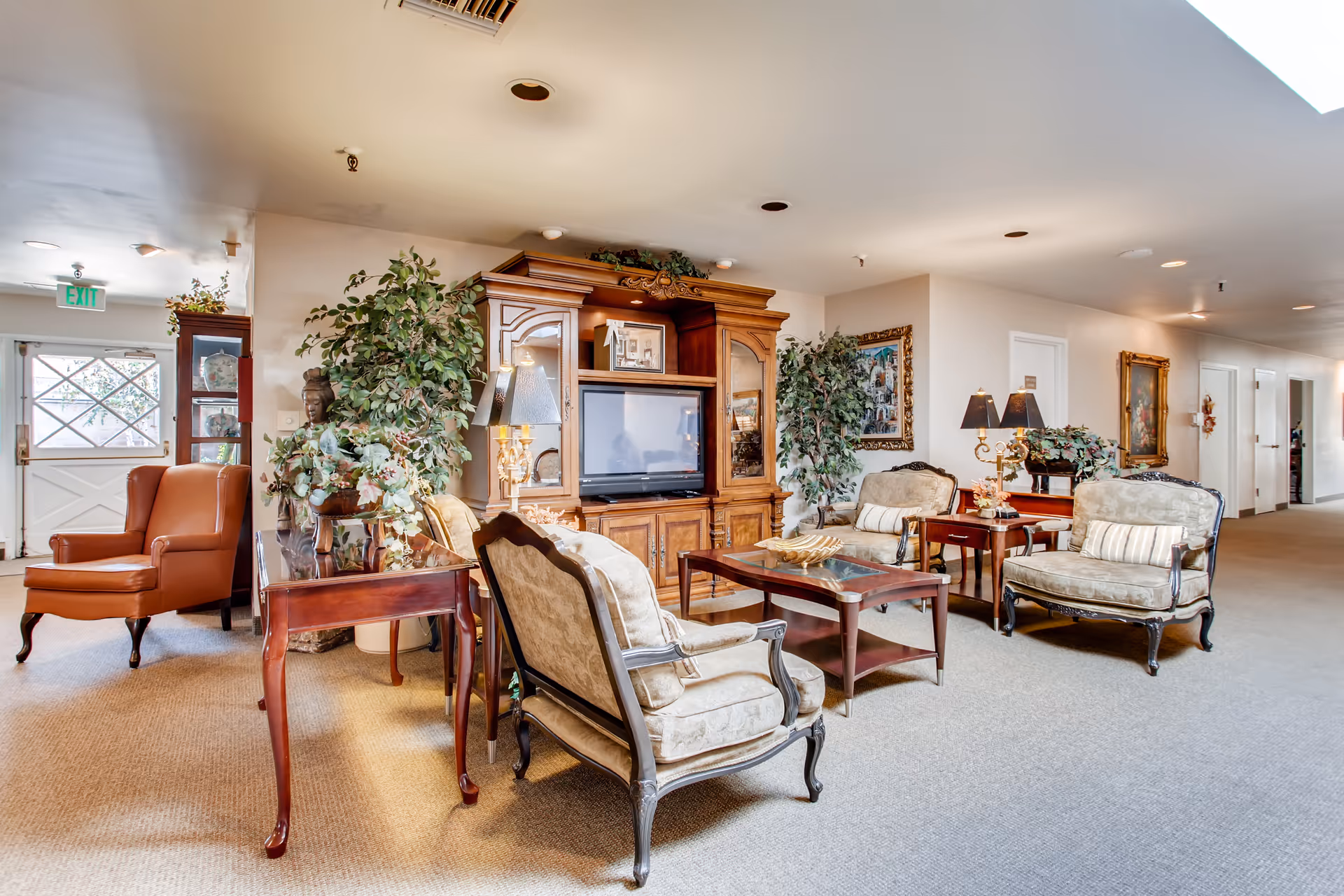 A cozy and well-lit living room area in a senior living facility featuring classic upholstered armchairs, a wooden coffee table, a large wooden entertainment center with a television, decorative plants, table lamps, and framed artwork on the walls. The room has a beige carpet and light-colored walls, with a hallway visible in the background.