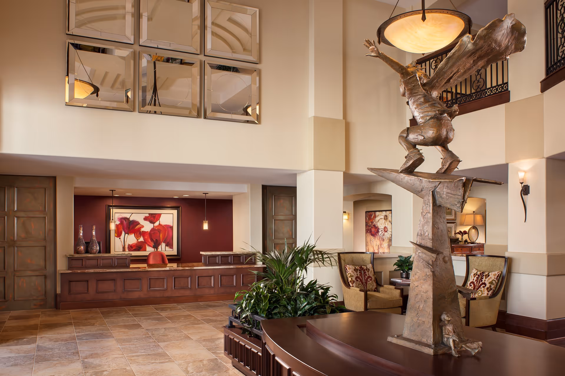 A spacious and elegant lobby area featuring a large bronze sculpture of a figure with wings on a pedestal. The lobby has a tiled floor, a wooden reception desk with decorative vases and a framed floral artwork behind it, comfortable seating with patterned cushions, indoor plants, and warm lighting fixtures including a hanging lamp and wall sconces.