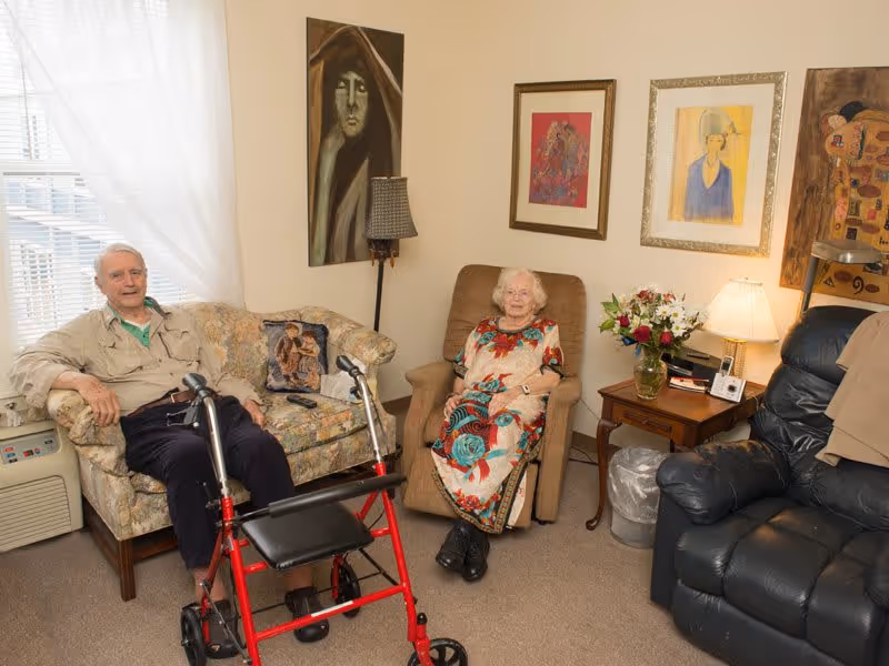 An elderly man and woman sitting in a cozy living room. The man is seated on a floral patterned couch with a red walker in front of him, and the woman is seated in a beige recliner chair. The room has several framed artworks on the walls, a side table with a lamp and a vase of flowers, and a black leather recliner chair. Light filters in through a window with sheer white curtains.