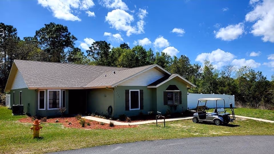 Front exterior of a single-story light green assisted living building with a golf cart parked on the lawn under a blue sky.