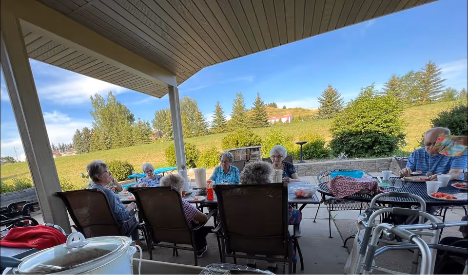 A group of elderly residents sitting and socializing around tables on a covered outdoor patio with walkers and a grassy hillside beyond.