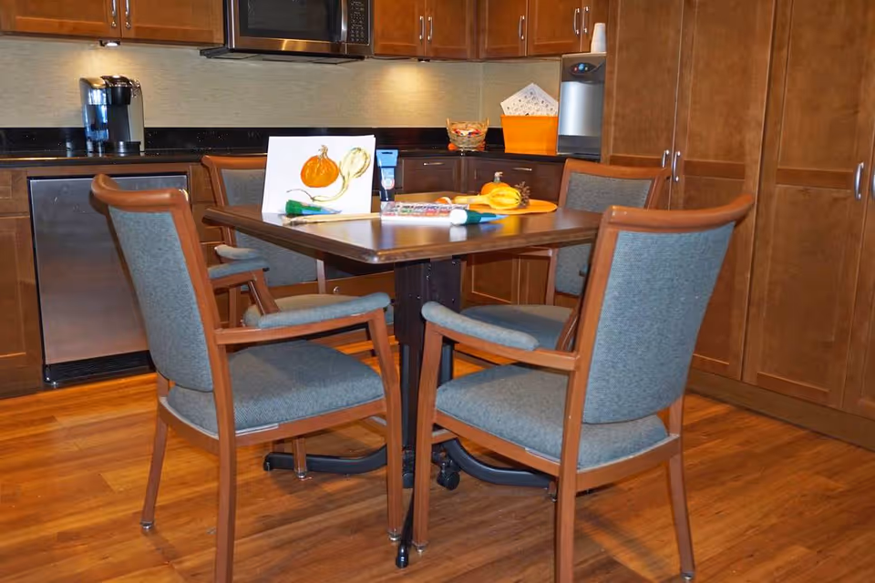 A kitchen area with wooden cabinets, a microwave, a coffee maker, and a small refrigerator. In the center, there is a wooden table with four cushioned chairs around it. On the table, there are some art supplies, a painting of pumpkins, and a small decorative plate with gourds.