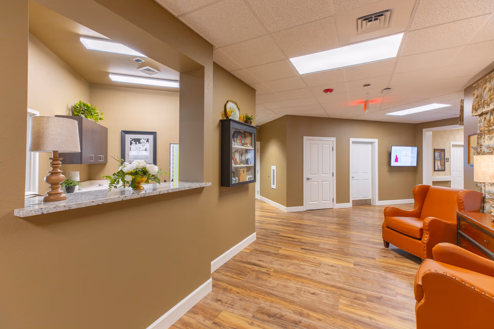 Interior view of a memory care community hallway with wood flooring, beige walls, and white doors. There is a reception window with a granite countertop displaying a lamp and a floral arrangement. Two orange leather armchairs are positioned near a wooden side table with a lamp. A wall-mounted TV and framed pictures are visible in the background.