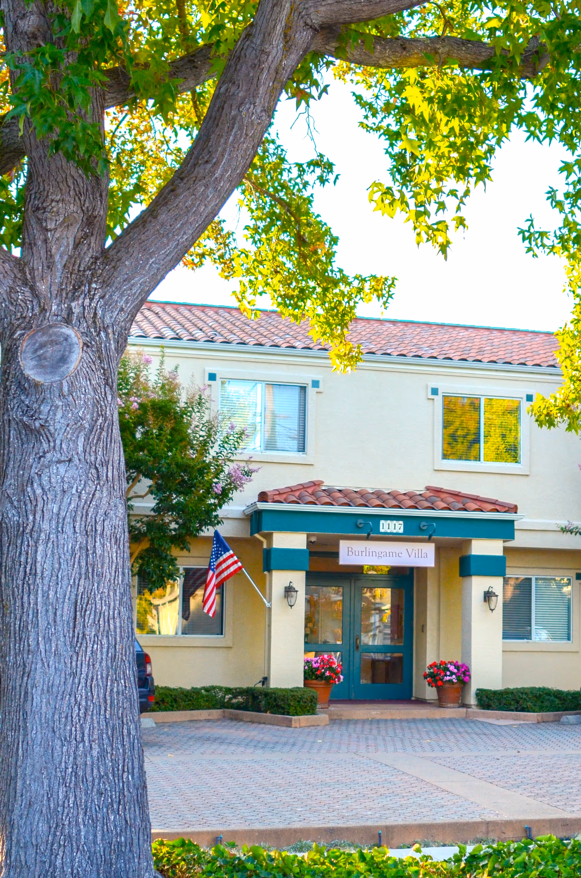 Front entrance of Burlingame Villa showing a tiled-roof facade, an American flag, potted flowers, and a large tree in the foreground.