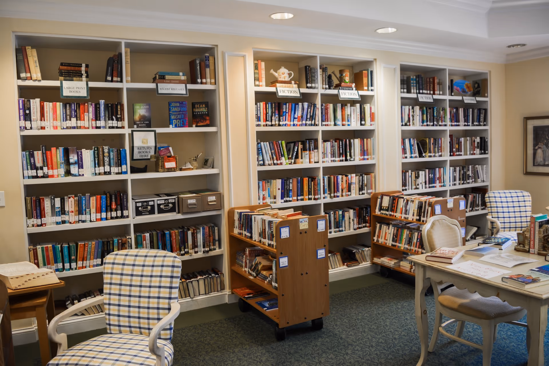 Interior view of a library room with white bookshelves filled with books labeled by categories such as Large Print Books and Fiction. There are two wooden book carts with additional books, a table with chairs, and a checkered upholstered armchair. The room has a cozy and organized atmosphere.