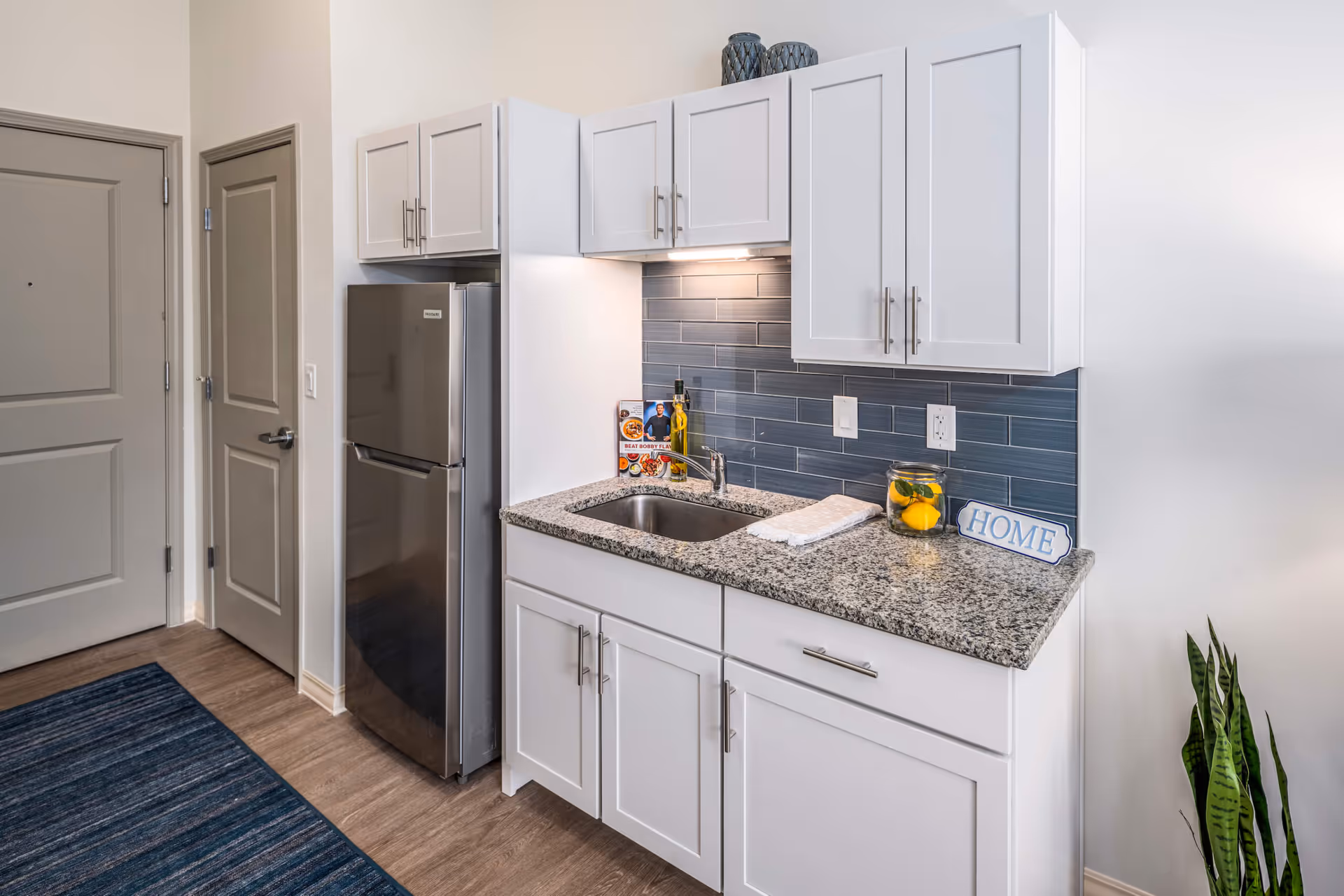A bright kitchenette with white cabinets, granite countertop, stainless-steel refrigerator, sink and blue tiled backsplash.