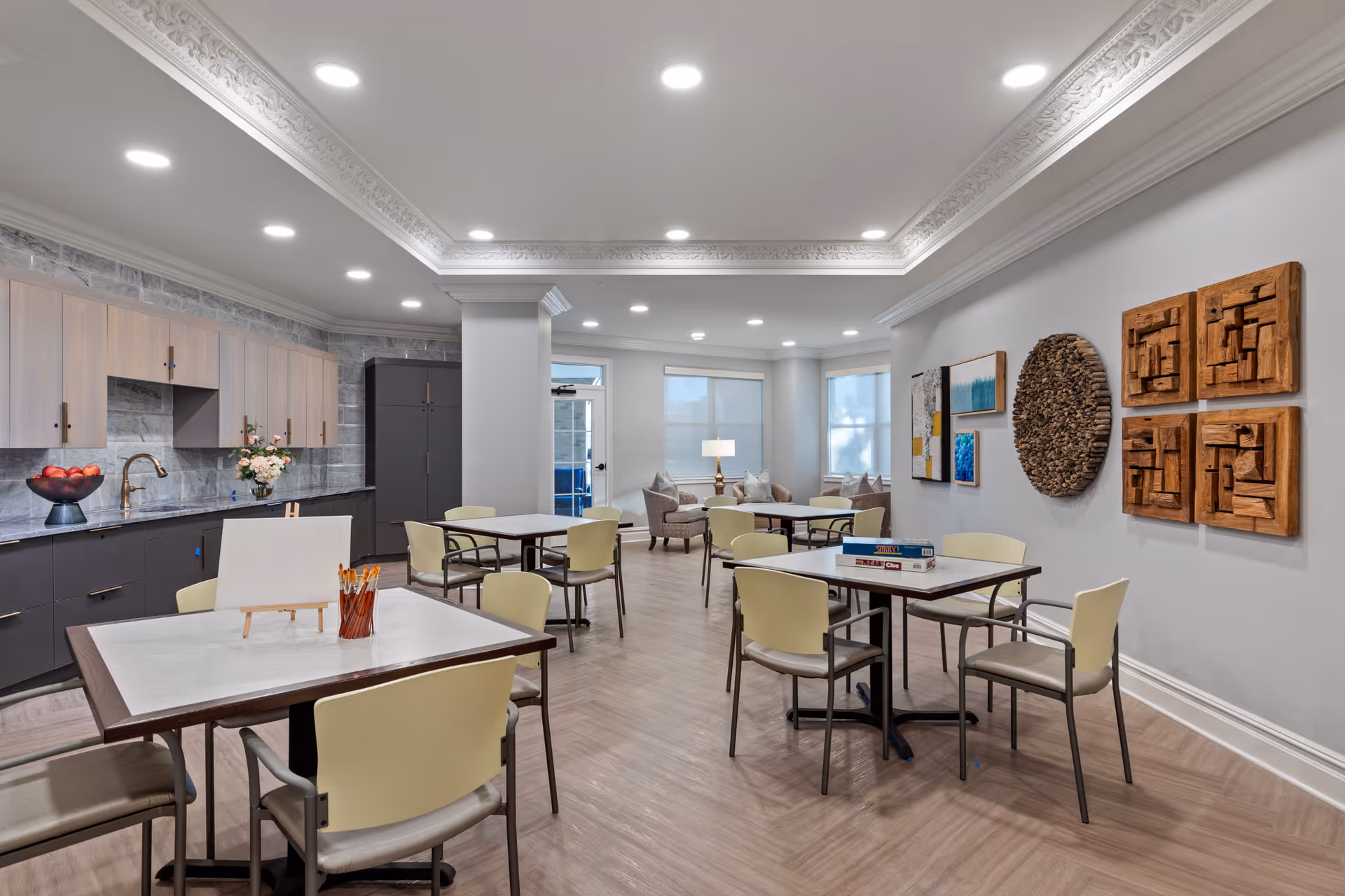 A well-lit common area with several tables and chairs arranged for group activities or dining. The room features a kitchenette with cabinets, a sink, and a bowl of apples on the counter. There are art pieces on the walls, including wooden sculptures and colorful paintings. In the background, a seating area with armchairs and a lamp is visible near large windows.