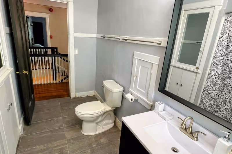 Bathroom interior with a toilet, sink and mirror, patterned shower curtain, and an open doorway to a hallway.