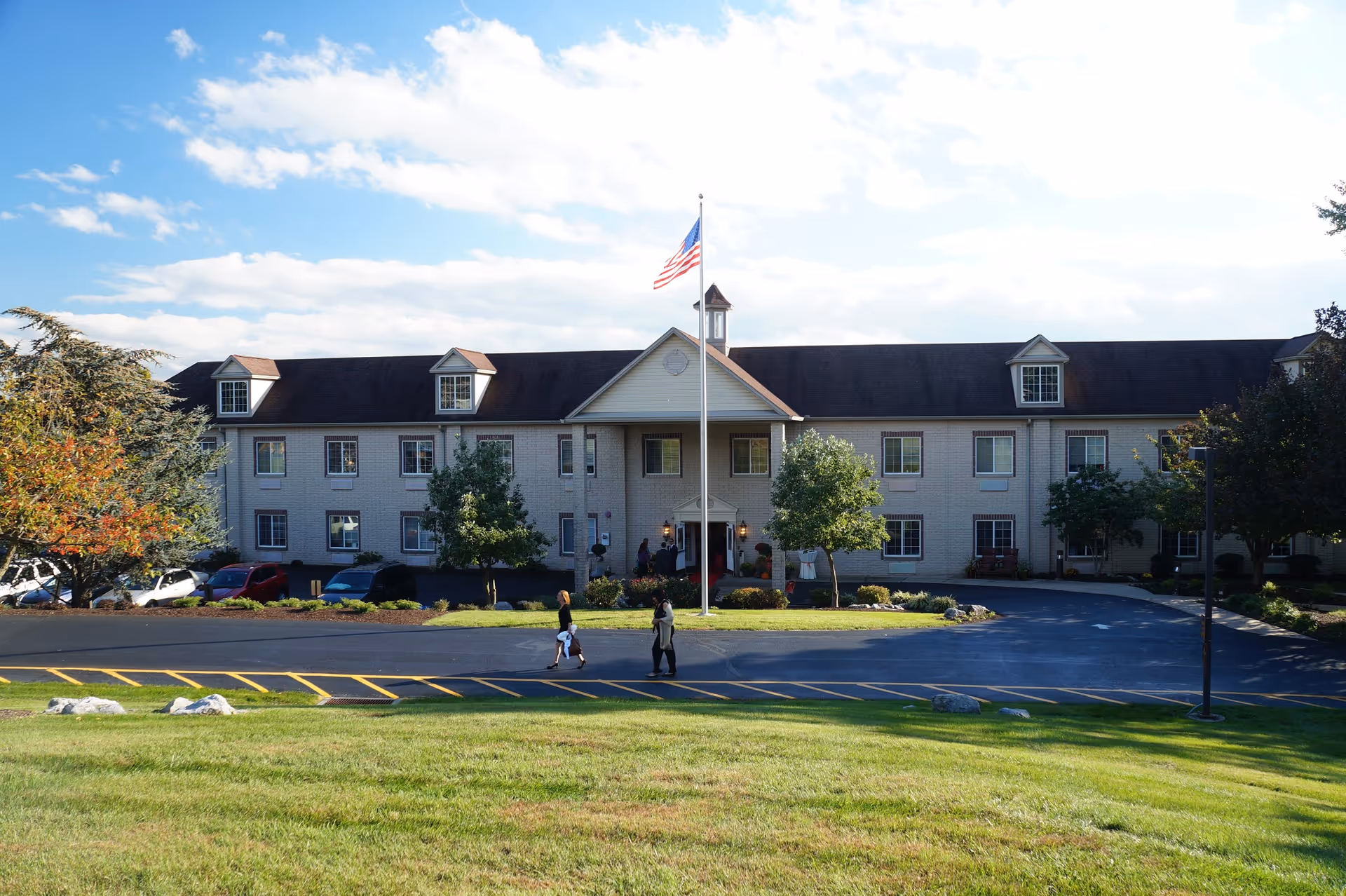 Front exterior of a two-story senior living building with an American flag on a flagpole, a lawn in the foreground, and two people walking toward the entrance.