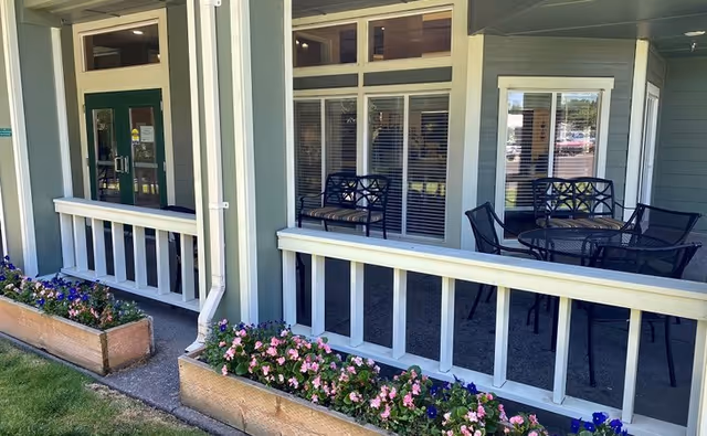 Covered outdoor patio area with white railings and flower boxes filled with pink and purple flowers. The patio has black metal chairs and tables, and large windows and doors leading inside the building.