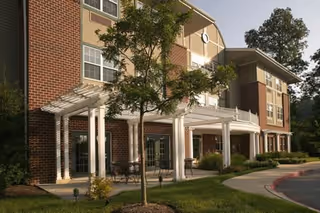 Front brick facade and covered entrance of a senior living facility with a porch, white columns, patio seating, and landscaped grounds.