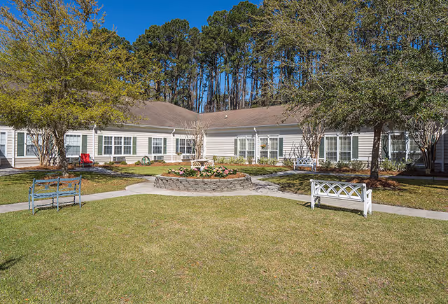 Outdoor courtyard area of a senior living facility with a circular flower bed in the center, surrounded by a paved walkway, benches, trees, and a single-story building with multiple windows in the background under a clear blue sky.