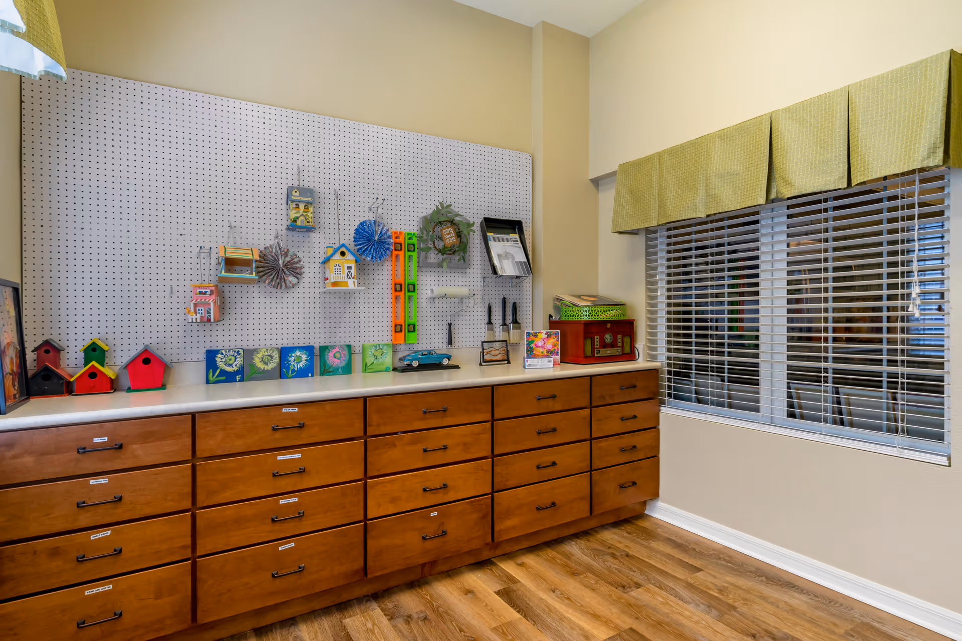 A craft room with a long wooden cabinet featuring multiple drawers, a pegboard on the wall displaying various craft items including small birdhouses, decorative flowers, and tools. There is a window with green valance curtains and wooden blinds, and the floor is wood.