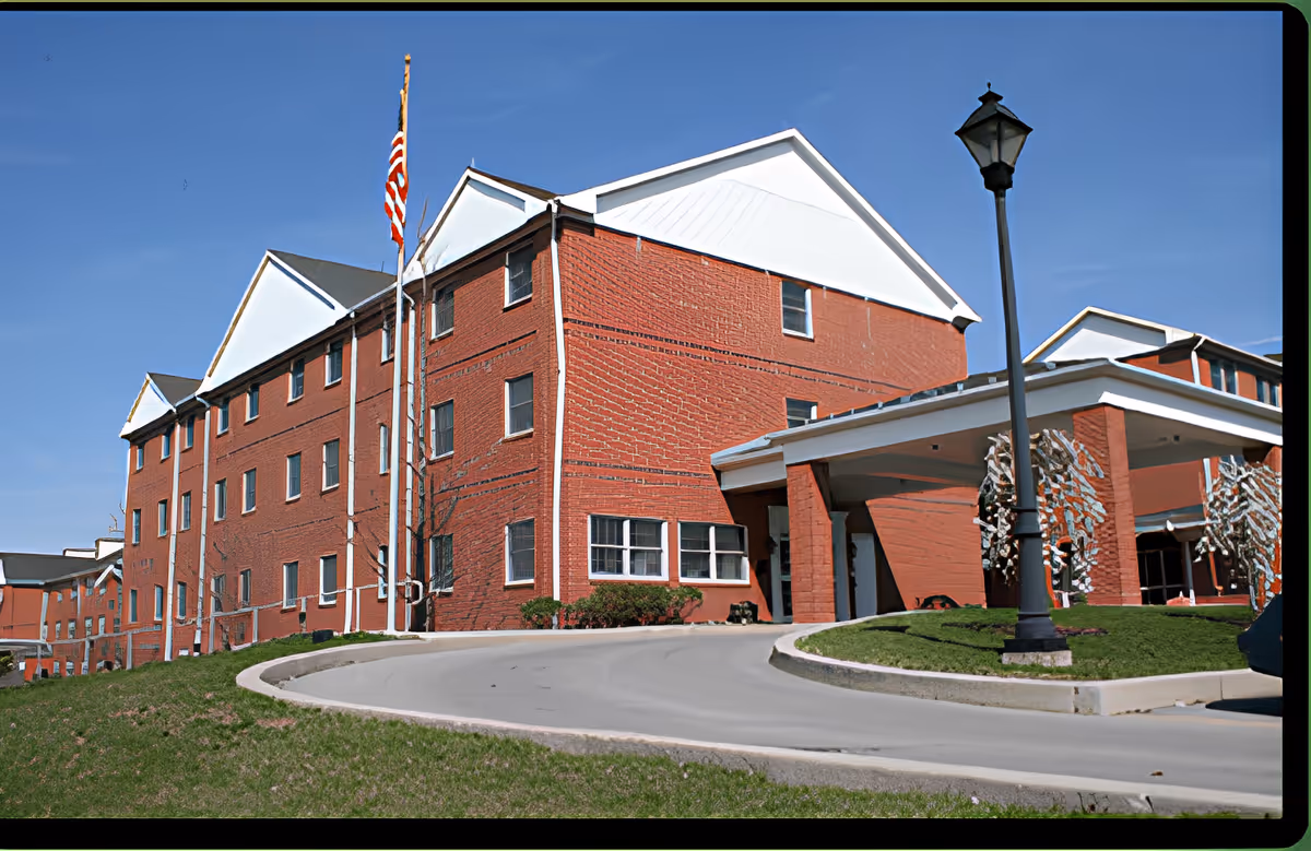 Exterior view of a large red brick building with white trim and multiple windows, featuring a covered entrance with columns, a flagpole with an American flag, and a street lamp on a curved driveway surrounded by grass under a clear blue sky.