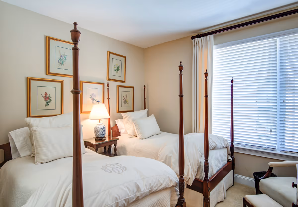 A cozy bedroom with two wooden four-poster twin beds dressed in white bedding. Between the beds is a wooden nightstand with a decorative lamp. The walls are adorned with framed botanical prints, and a large window with white blinds and cream curtains lets in natural light. A cushioned chair and ottoman are positioned near the window.