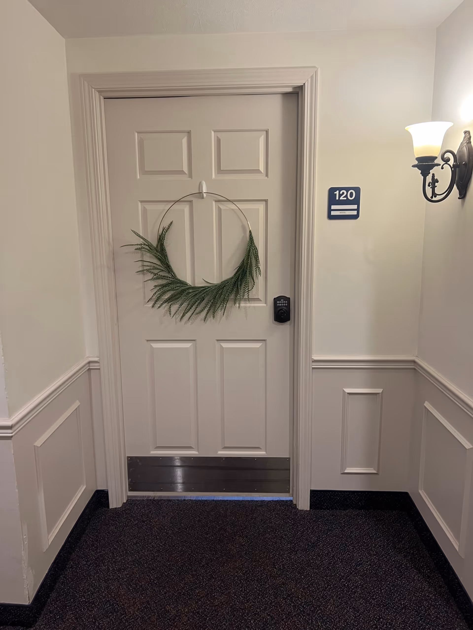 A closed beige door with a decorative green wreath hanging on it, labeled with the number 120. The door has a digital keypad lock and a metal kick plate at the bottom. The surrounding walls are painted in light colors with decorative molding, and there is a wall-mounted light fixture on the right side.