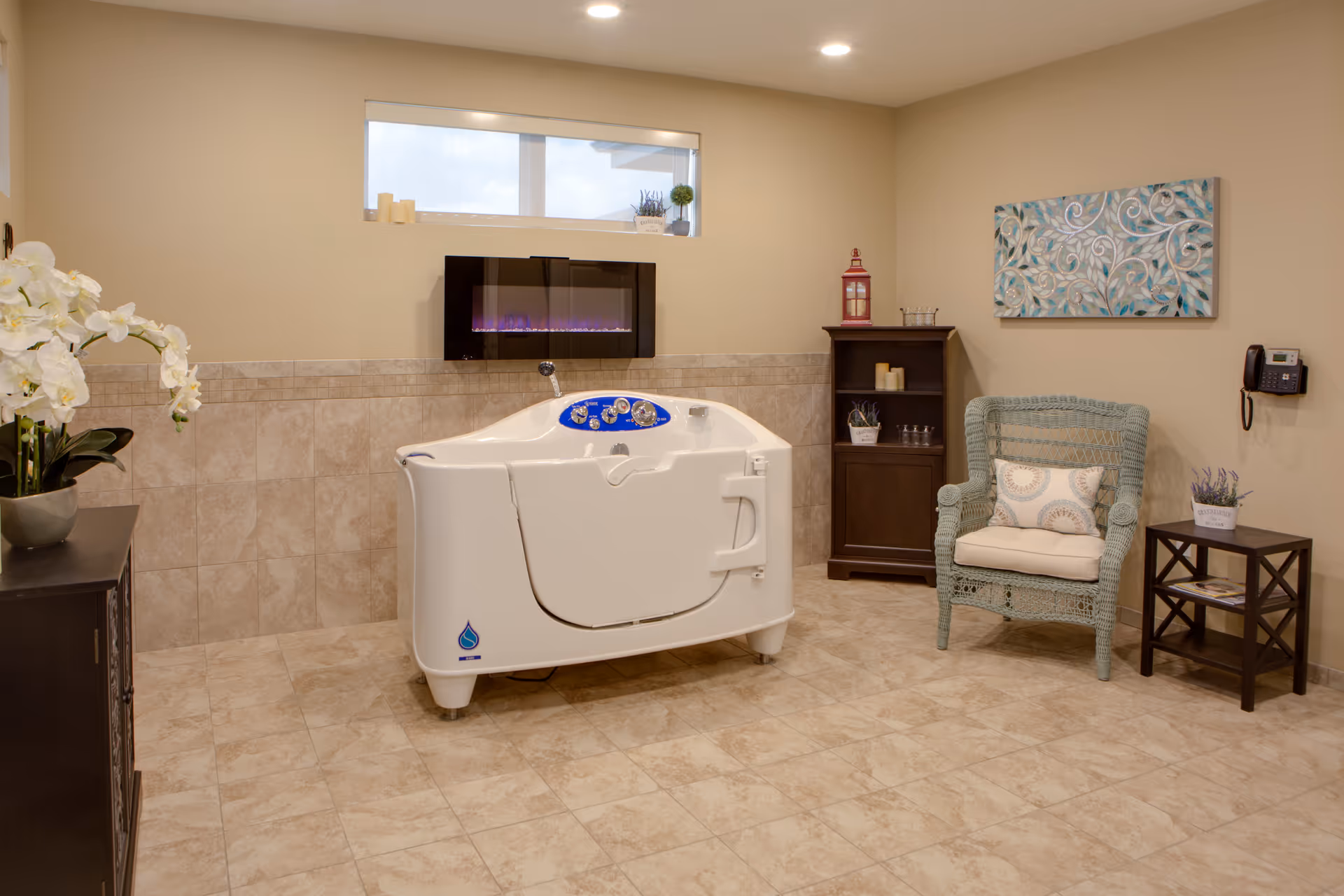 A spacious, well-lit bathroom featuring a white walk-in bathtub with control knobs, beige tiled walls and floor, a small window above the tub, a modern electric fireplace mounted on the wall, a wicker armchair with a cushion, a small wooden side table with a potted plant, a dark wooden cabinet with decorative items, and a wall-mounted phone.