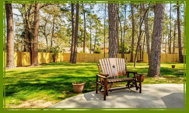 A wooden bench with a star design on the backrest sits on a concrete patio in a fenced backyard surrounded by tall pine trees and green grass under a clear sky.