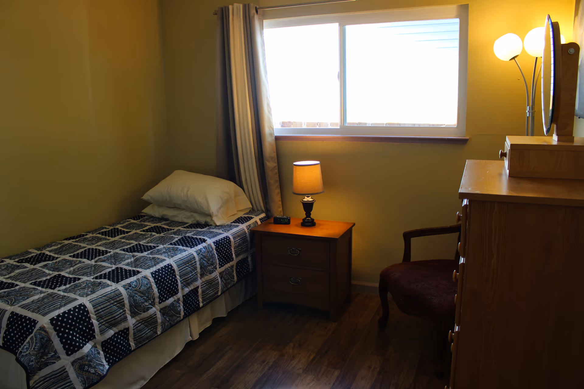 A small bedroom with a single bed covered with a patterned quilt and a pillow. Next to the bed is a wooden nightstand with a lamp and a digital clock. There is a window with curtains letting in natural light. On the right side, there is a wooden dresser with a mirror and a floor lamp beside a cushioned chair.