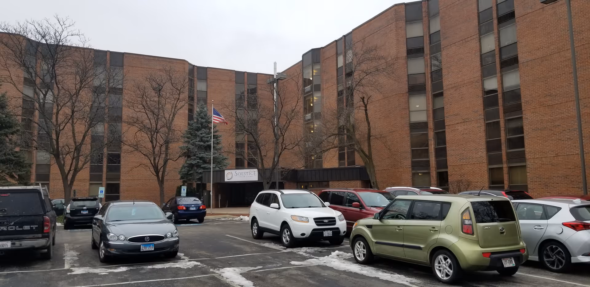 Exterior view of a multi-story brick building with several windows, a parking lot with multiple parked cars, leafless trees, and an American flag on a flagpole near the entrance. A sign near the entrance reads 'Solstice'.