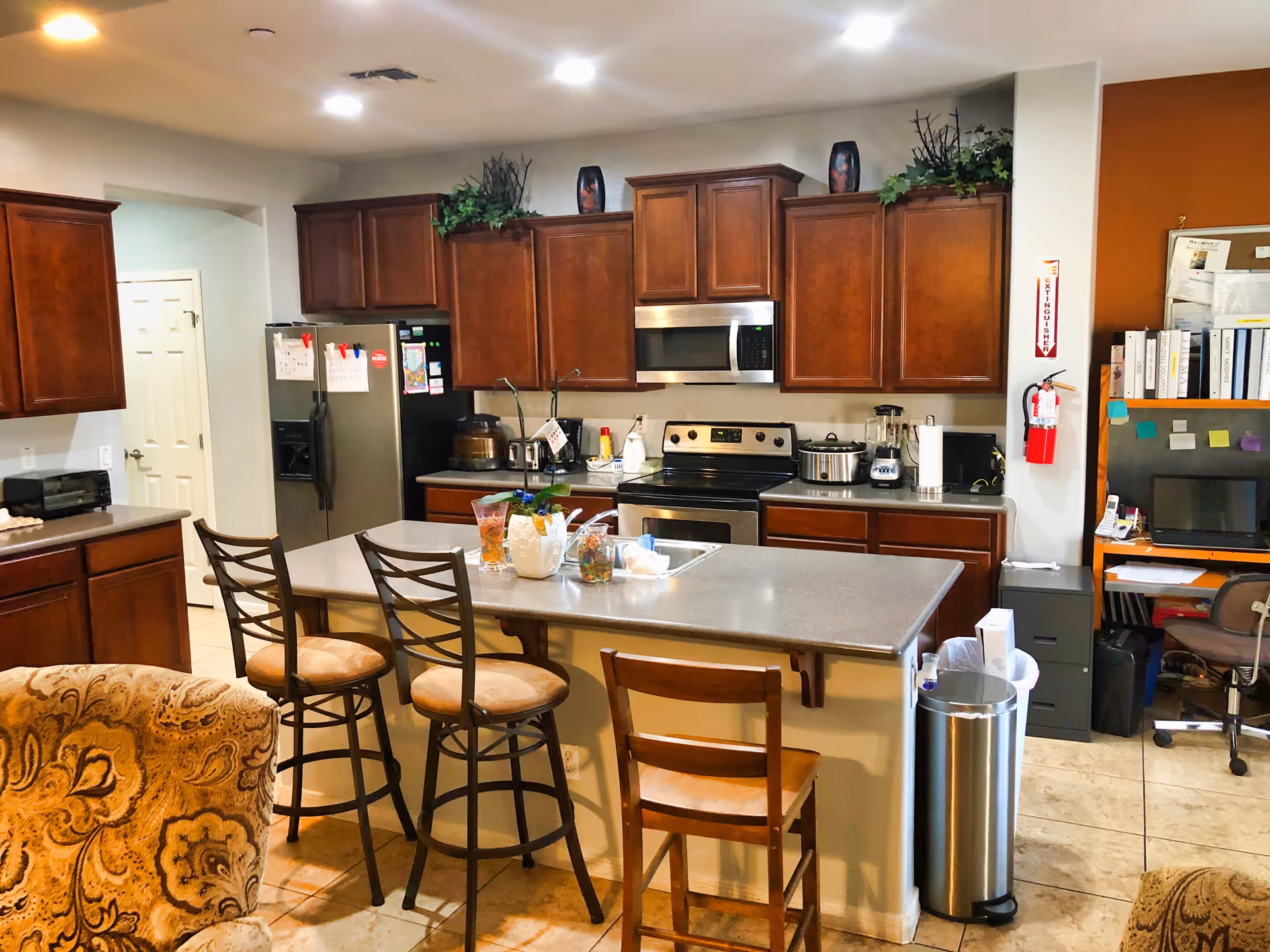A well-lit kitchen area with wooden cabinets, a stainless steel refrigerator, stove, microwave, and various kitchen appliances on the countertops. There is a kitchen island with three chairs, two metal stools with cushioned seats and one wooden chair. A fire extinguisher is mounted on the wall next to a small office area with a desk, computer, and shelves filled with binders and papers.