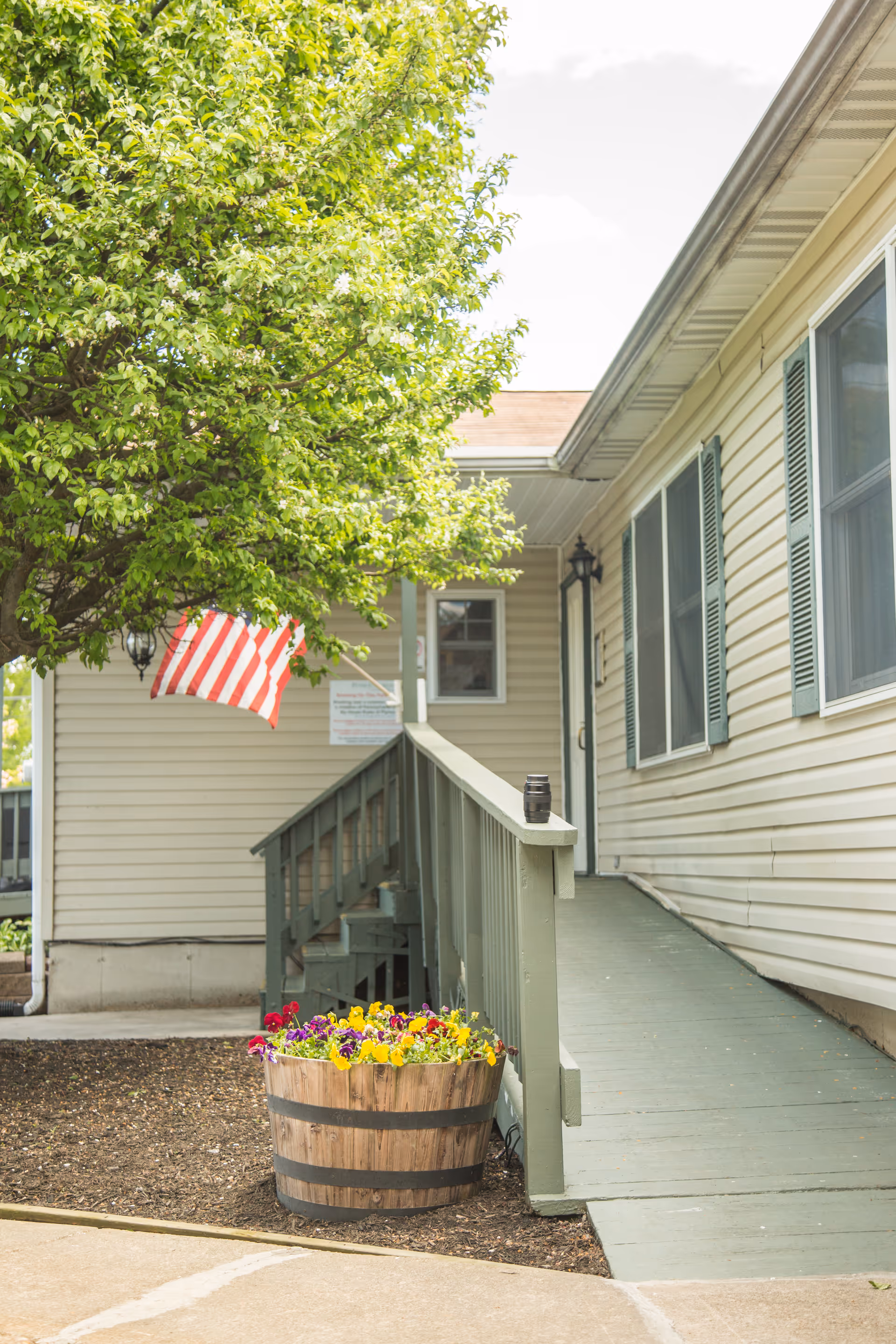 Exterior view of a beige-sided building entrance with a green ramp, American flag, tree, and a wooden flower planter.