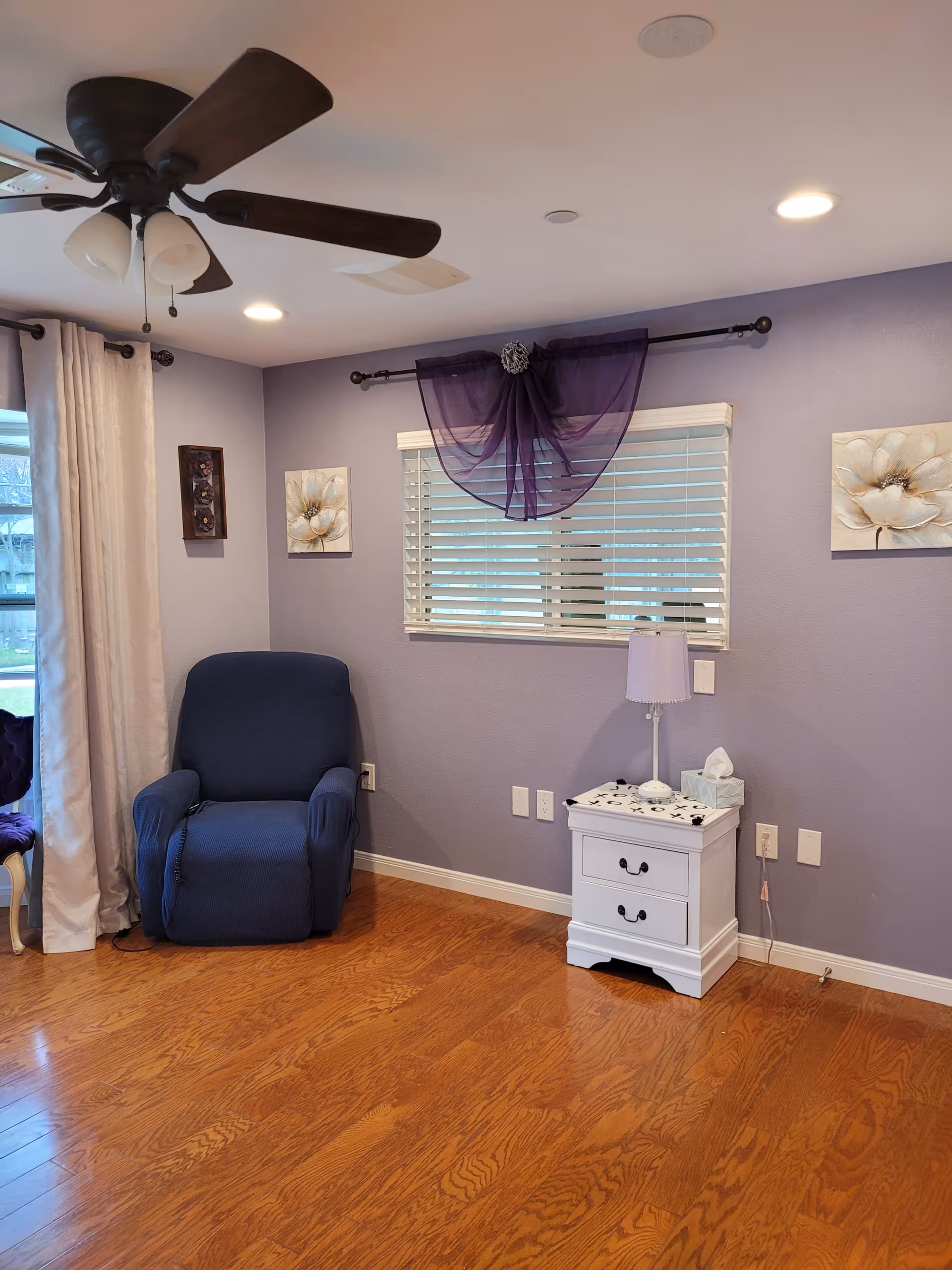 A cozy room with light purple walls and wooden flooring. There is a dark blue recliner chair next to a window with white blinds and a sheer purple curtain swag. A white nightstand with two drawers holds a small lamp and a tissue box. The room is decorated with two floral paintings on the walls and has a ceiling fan with dark wooden blades and three light fixtures.