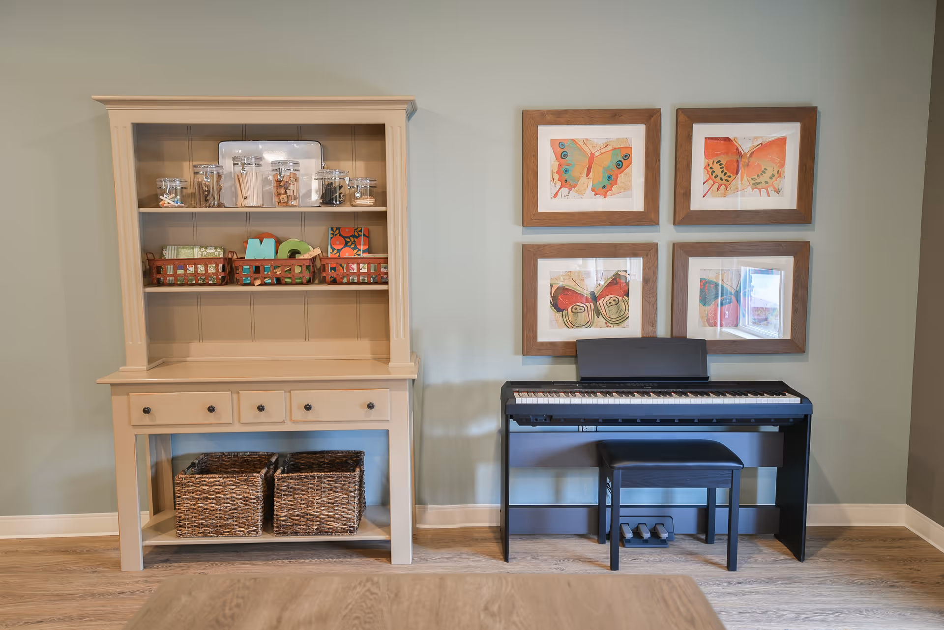 A cozy interior corner featuring a beige wooden cabinet with shelves holding jars and baskets, next to a black digital piano with a matching bench. Above the piano, there are four framed butterfly artworks arranged in a grid on a light green wall. The floor is wooden.
