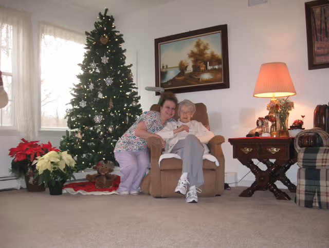 A younger woman kneels beside an elderly woman seated in an armchair next to a decorated Christmas tree in a cozy living room.