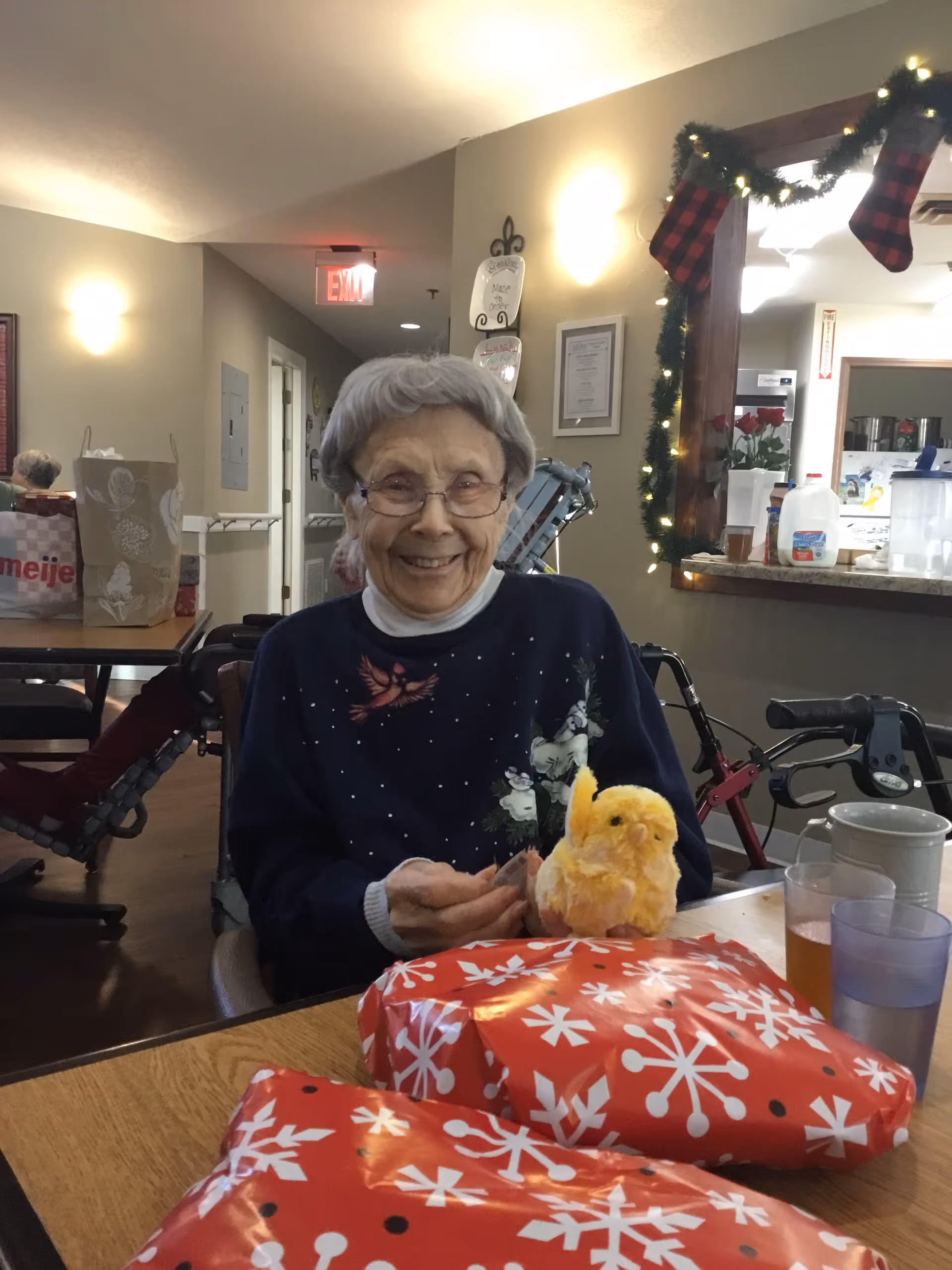 An elderly woman with gray hair and glasses is sitting at a table in a communal area, smiling. She is wearing a dark sweater with a cardinal bird and snowman design. On the table in front of her are two red and white snowflake-patterned wrapped gifts, a yellow plush chick, a glass of iced tea, and a cup. The background shows a decorated window frame with Christmas stockings and garland, a walker, and other tables with chairs in a well-lit room.