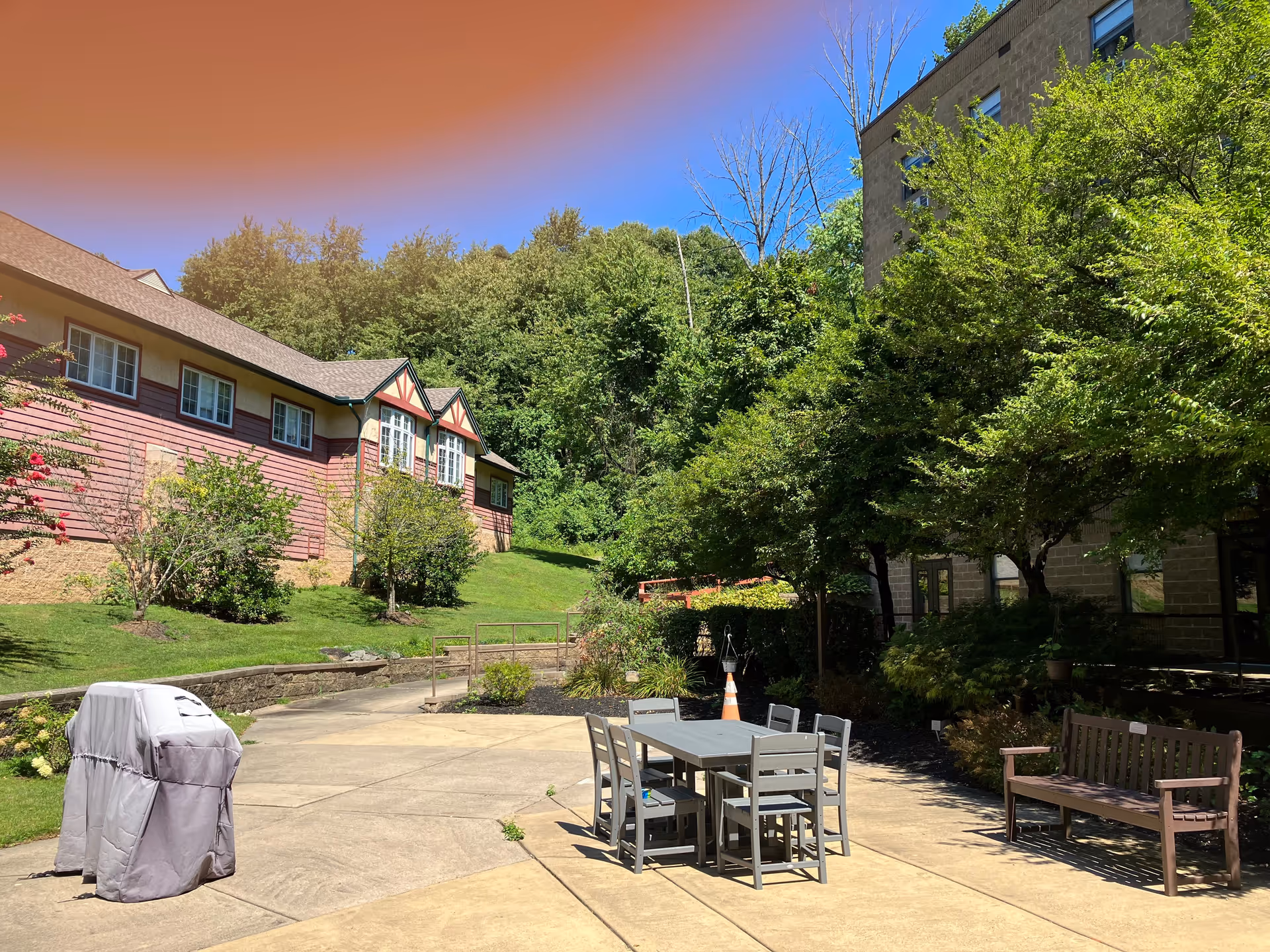 Outdoor patio area at Gloria Dei Manor featuring a covered grill, a table with six chairs, a wooden bench, surrounded by trees and greenery with buildings in the background under a clear blue sky.