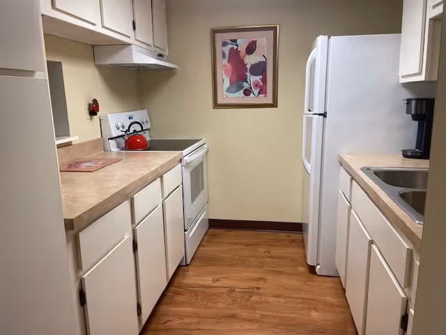 A small kitchen with white cabinets and beige countertops on both sides. On the left side, there is a white stove with a red kettle on top and a range hood above it. On the right side, there is a white refrigerator and a sink with a coffee maker next to it. The floor is wooden, and a framed floral painting hangs on the wall at the end of the kitchen.