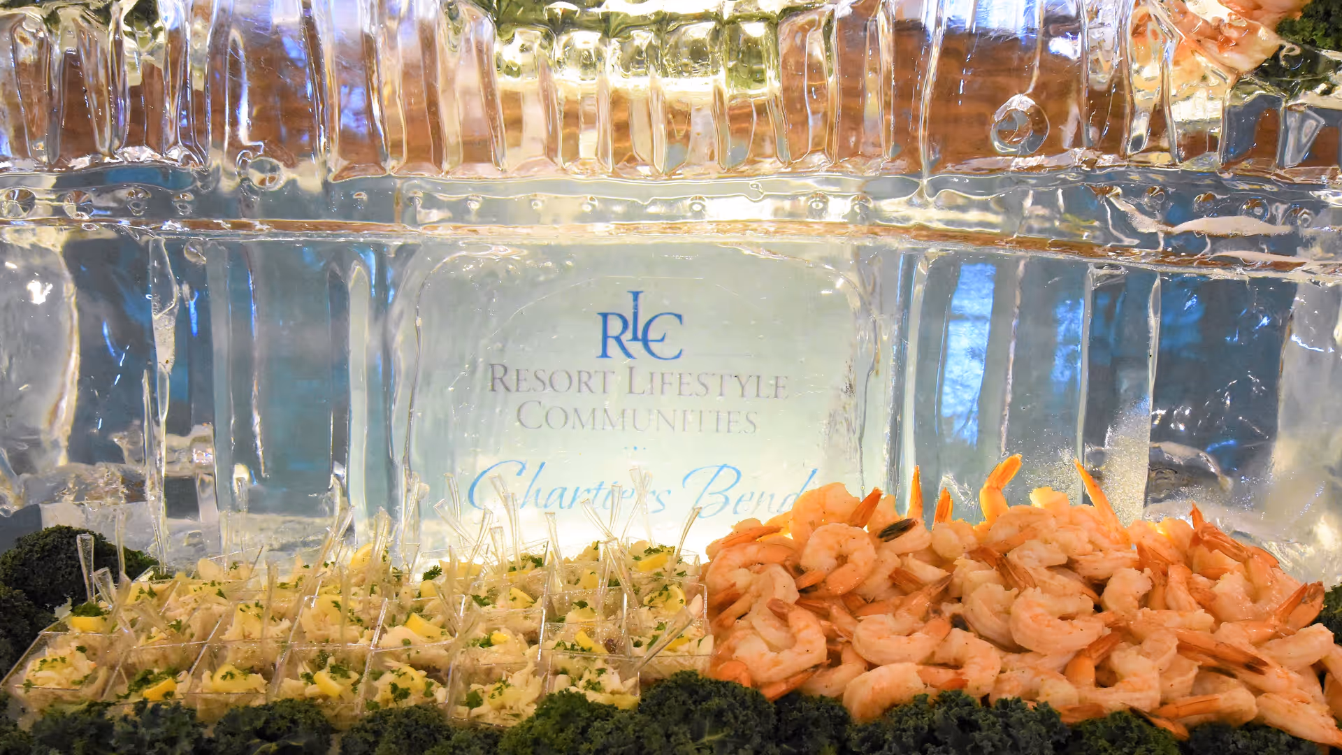 A seafood buffet display featuring a large pile of cooked shrimp on the right and individual servings of a salad or seafood dish with toothpicks on the left, all arranged in front of an ice sculpture with the text 'Resort Lifestyle Communities Chartiers Bend'.