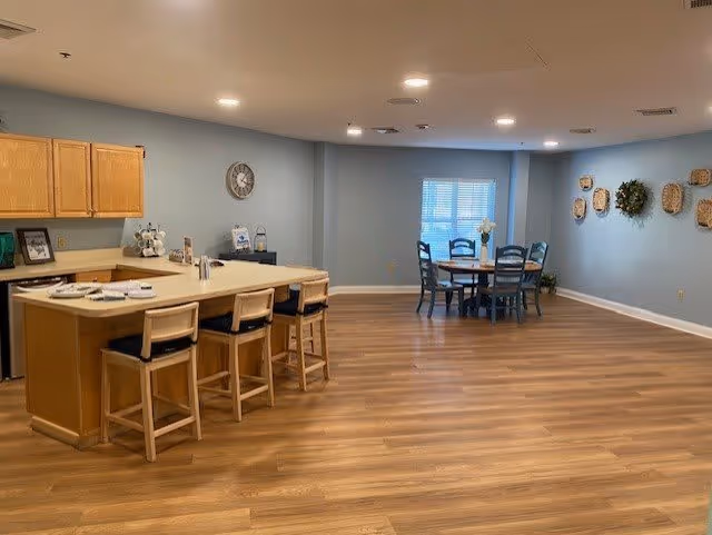 A spacious room with a kitchen area featuring wooden cabinets and a countertop with four wooden bar stools. In the background, there is a round dining table with four chairs near a window with blinds. The walls are painted light blue and decorated with a clock and wall hangings. The floor is covered with wood-style flooring and the ceiling has recessed lighting.