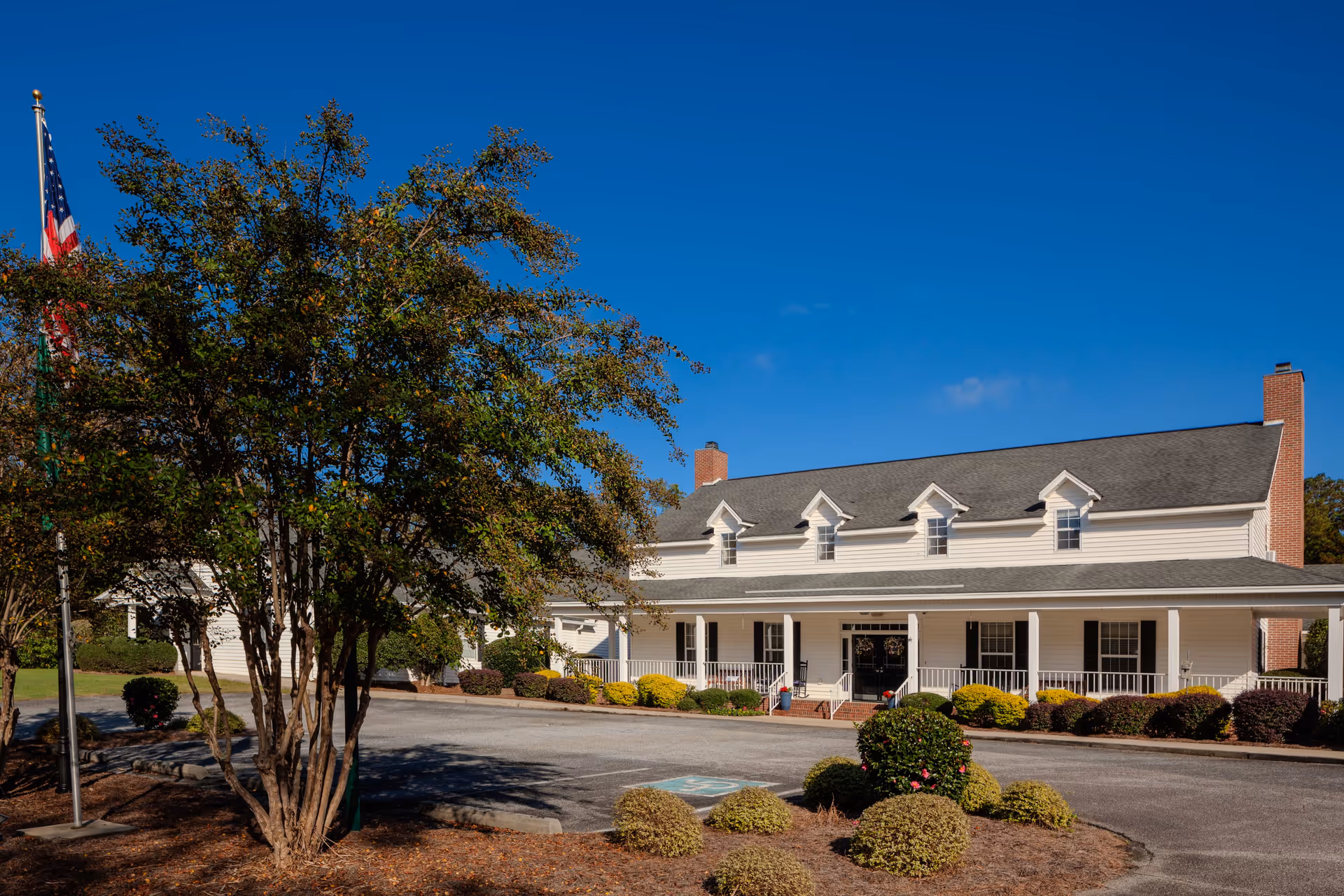 Front exterior of a two-story senior living building with a covered porch, landscaped shrubs, and an American flag.