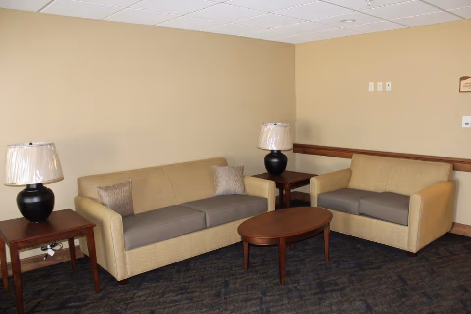 A small sitting area with two beige sofas, each with two cushions, positioned around a wooden oval coffee table. There are two wooden side tables, each with a black lamp with a white lampshade. The walls are beige and the floor is carpeted in a dark pattern.
