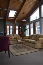 Sunlit living room with a vaulted wood ceiling and skylights, large windows, two sofas, a coffee table and an area rug.