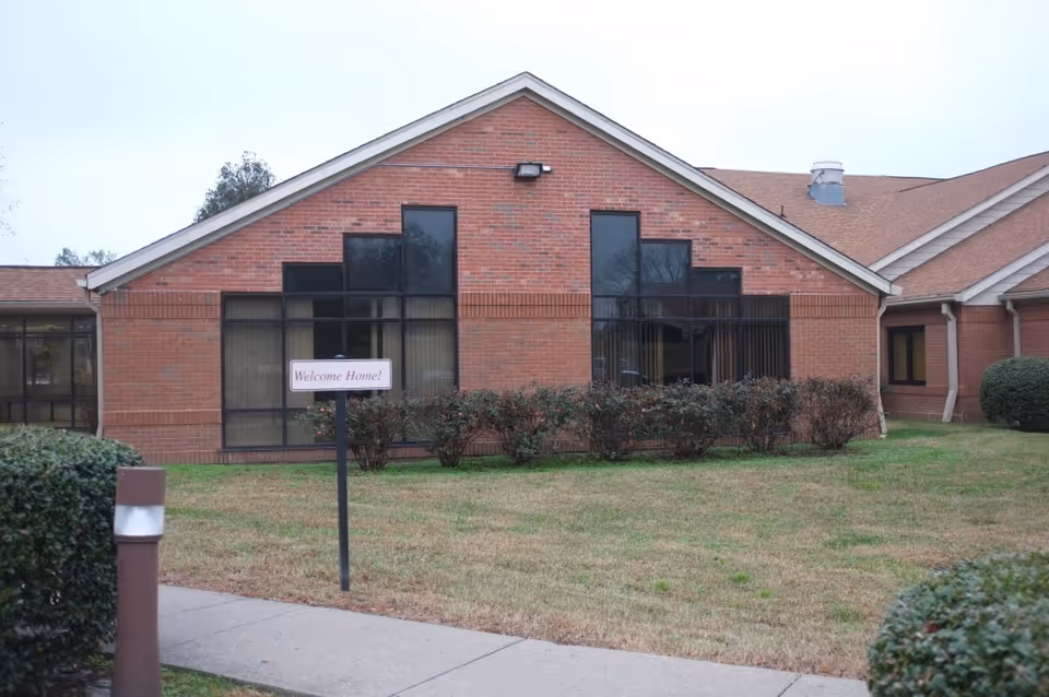Exterior view of a brick building with large windows and a gable roof. In front of the building, there is a small garden with bushes and a sign that reads 'Welcome Home!'. A sidewalk and some trimmed bushes are visible in the foreground.