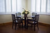 A round wooden dining table with a floral centerpiece surrounded by four wooden chairs with cushioned seats, set in a room with large windows covered by white plantation shutters and wooden flooring.