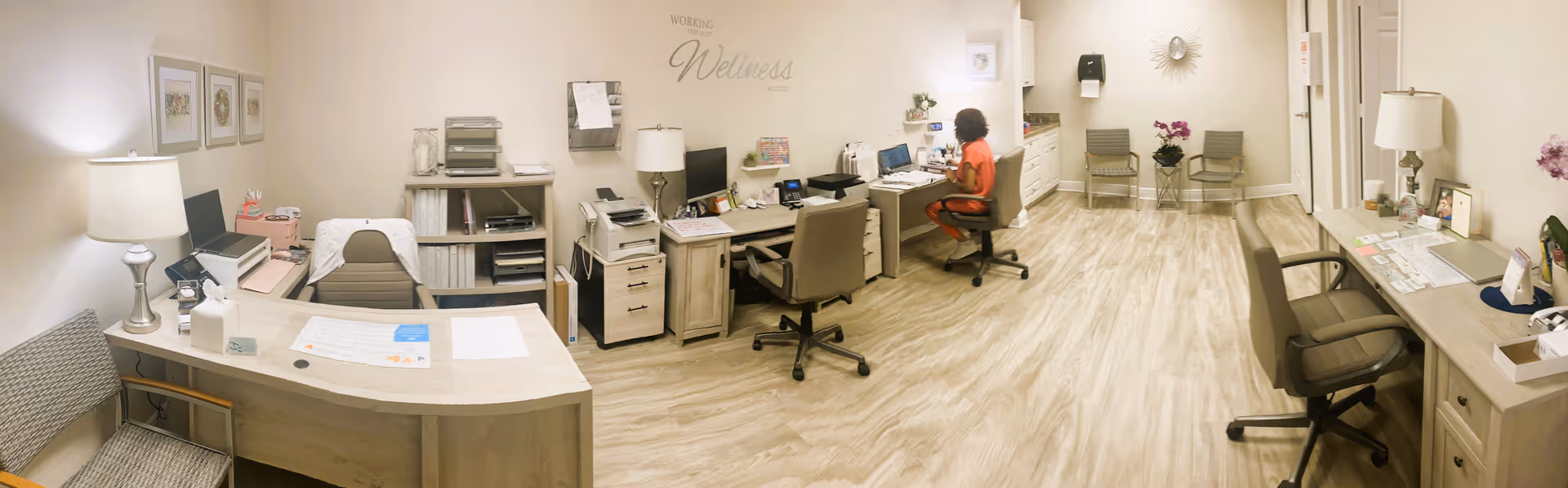 A spacious office area with multiple desks and chairs arranged along the walls. The room has light-colored wooden flooring and soft beige walls. On the left side, there is a desk with a lamp, printer, and office supplies. In the center, a person wearing an orange shirt is seated at a desk working on a computer. The wall behind them has the words 'Working Wellness' displayed. The right side features more desks with office equipment, chairs, and decorative items including a lamp and flowers. The room is well-lit with a calm and organized atmosphere.